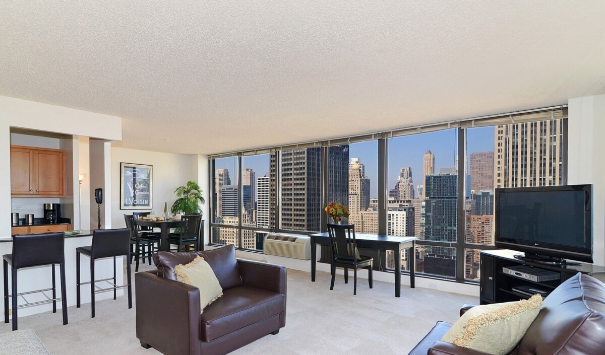 Modern apartment living room with large windows offering a clear city skyline view, furnished with chairs, a TV, dining table, and kitchen area.