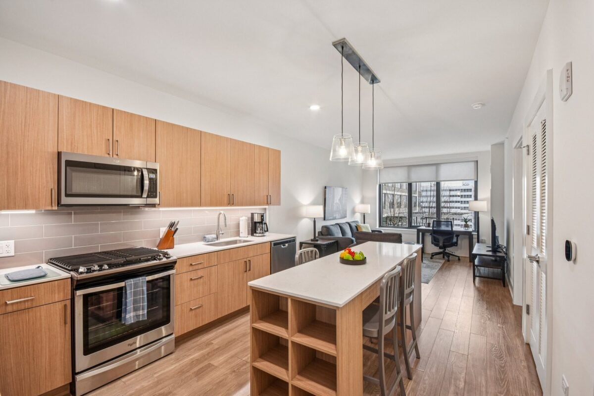 Modern kitchen with wood cabinets, stainless steel appliances, an island with bar stools, and an open view into a bright living area with large windows.