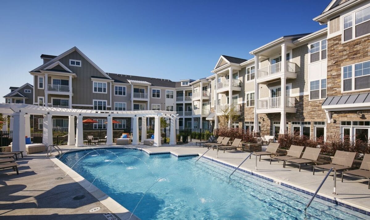 Outdoor swimming pool area with lounge chairs, sunshades, and a pergola, surrounded by modern multi-story apartment buildings under a clear blue sky.