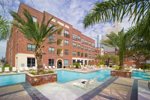 A brick apartment building with balconies overlooks a large outdoor swimming pool surrounded by lounge chairs and palm trees, with city skyscrapers visible in the background.