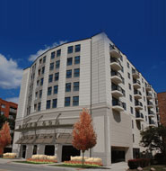 A six-story modern apartment building with balconies and large windows, set against a clear blue sky with trees in front.