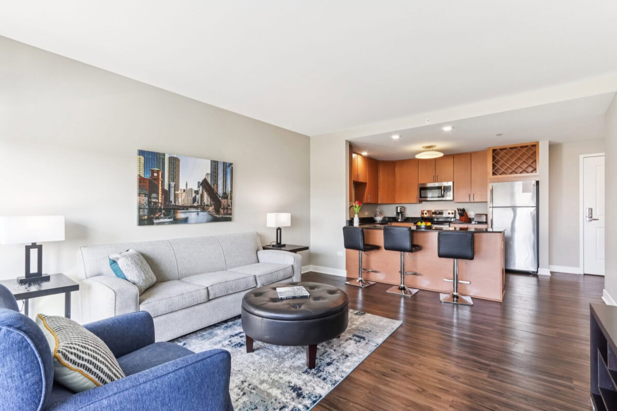 Modern apartment living area with a gray sofa, blue armchair, round ottoman, open kitchen with bar stools, and stainless steel appliances.