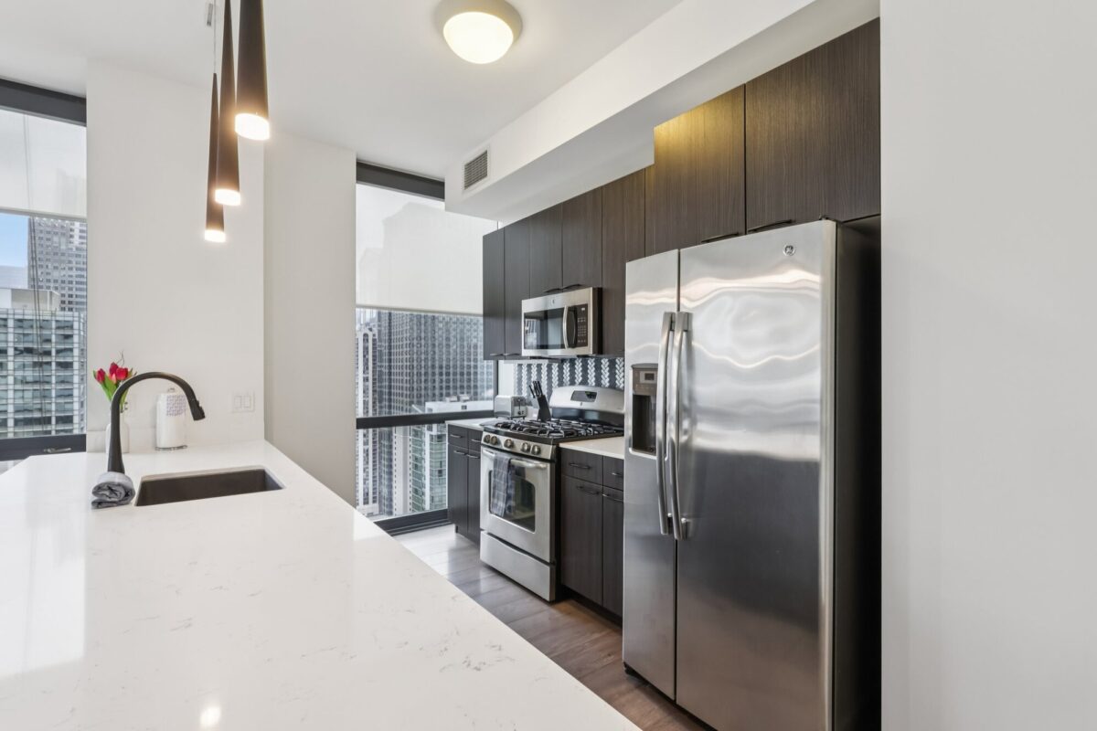 Modern kitchen with stainless steel appliances, dark cabinets, white quartz countertops, pendant lights, and large windows overlooking a cityscape.