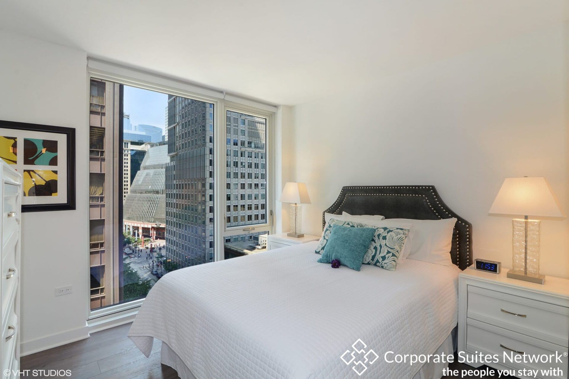 A modern bedroom with a large window showing city buildings, white bedding, nightstands with lamps, and decorative pillows on the bed.