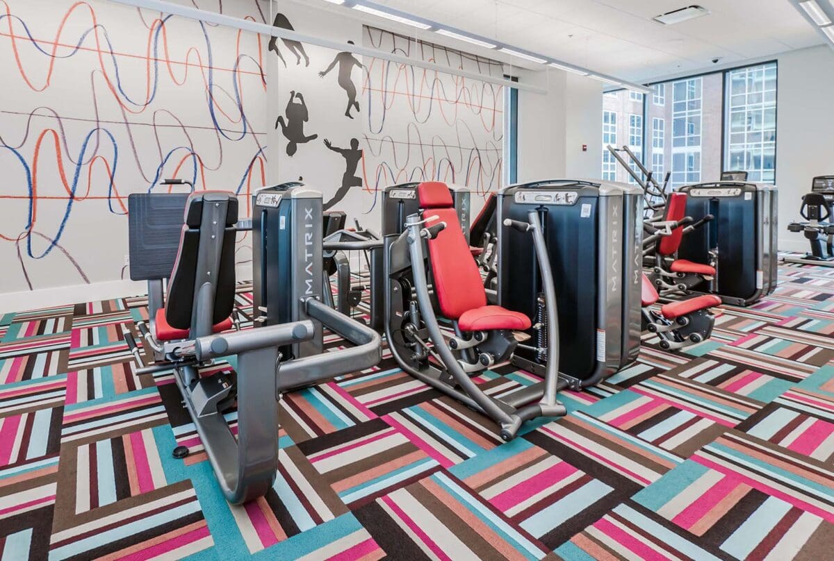 A gym room with brightly colored patterned carpet, red and black exercise machines, large windows, and a wall mural featuring abstract lines and silhouettes of athletes.