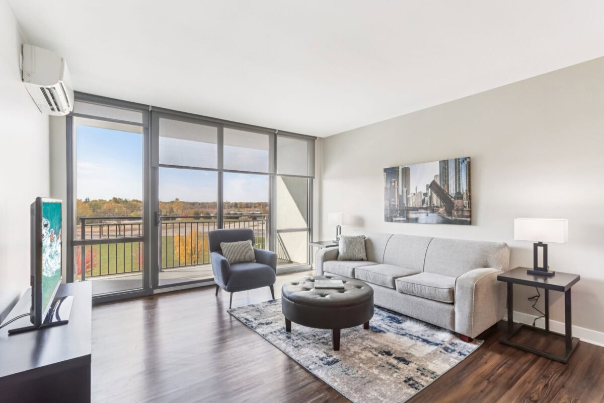 Modern living room with large windows, gray sofa, armchair, round ottoman, TV, and cityscape artwork, featuring wood floors and a balcony view.