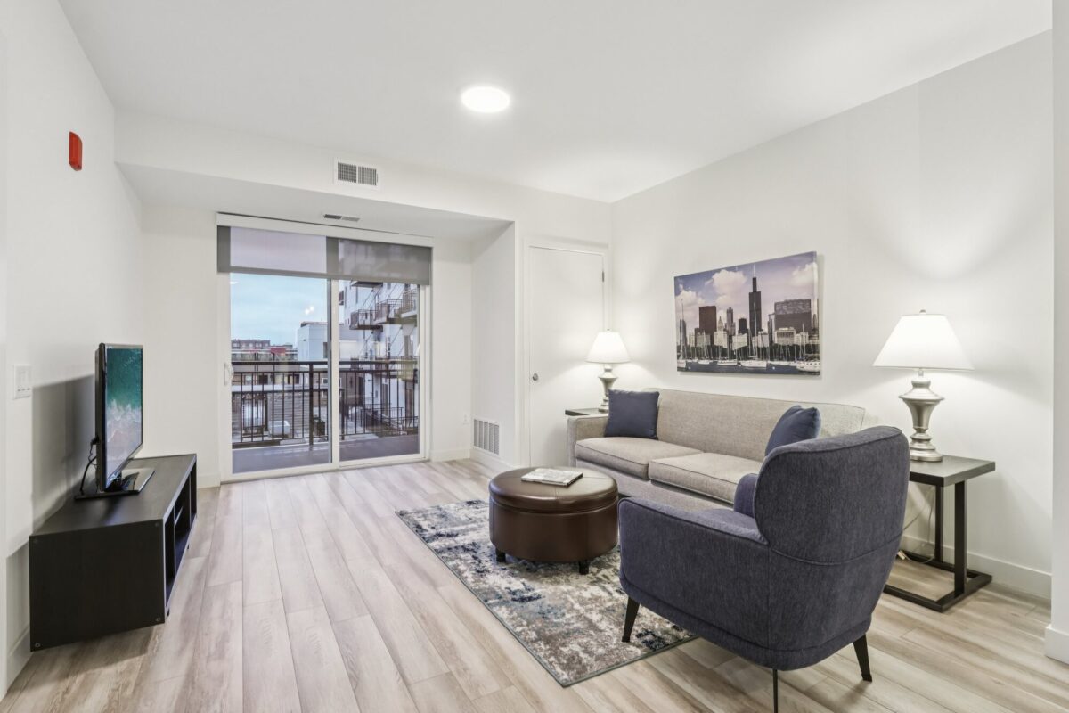 Modern living room with light wood floors, gray furniture, a TV on a stand, side tables with lamps, artwork on the wall, and a sliding glass door leading to a balcony.