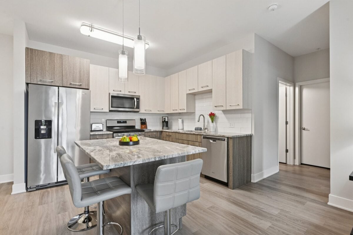 Modern kitchen with stainless steel appliances, marble island with two gray chairs, light wood cabinets, and a bowl of fruit on the counter.