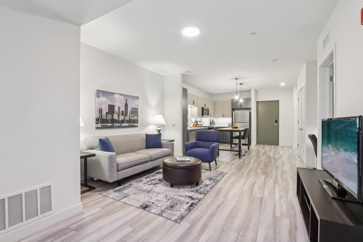 Modern open-concept living room and kitchen with light wood floors, gray sofa, blue chair, wall art, TV, and stainless steel appliances.