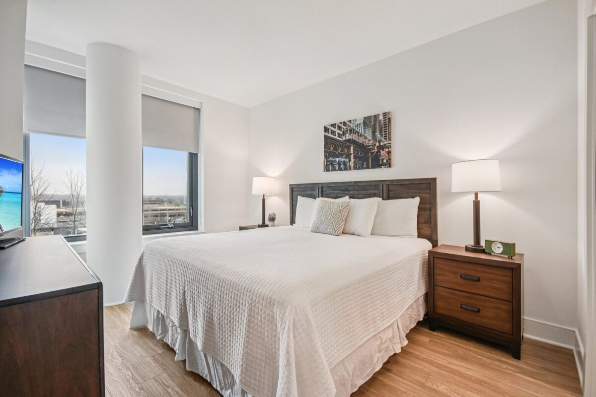 A bright bedroom with a large bed, two bedside tables with lamps, a dresser with a TV, light wood flooring, and a window with a city and water view.