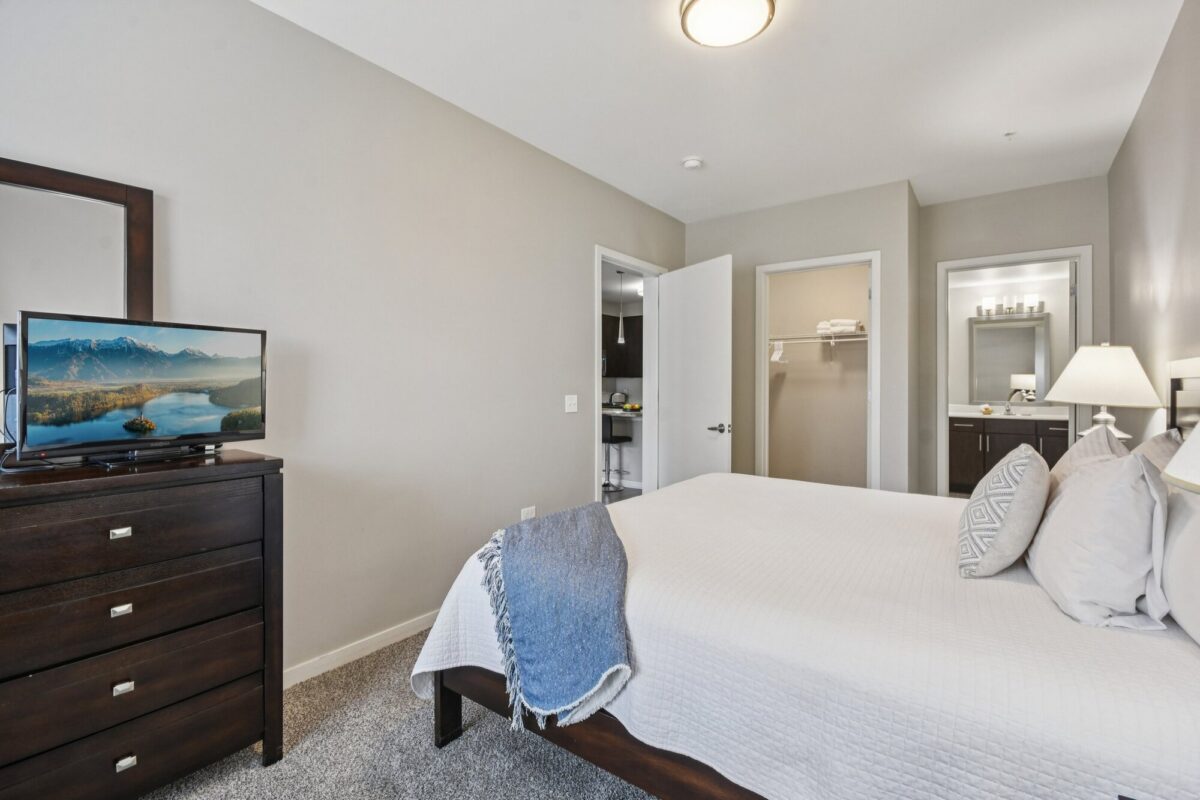 A neatly made bed with white linens in a modern bedroom, a TV on a dresser, and open doors leading to a bathroom and closet.