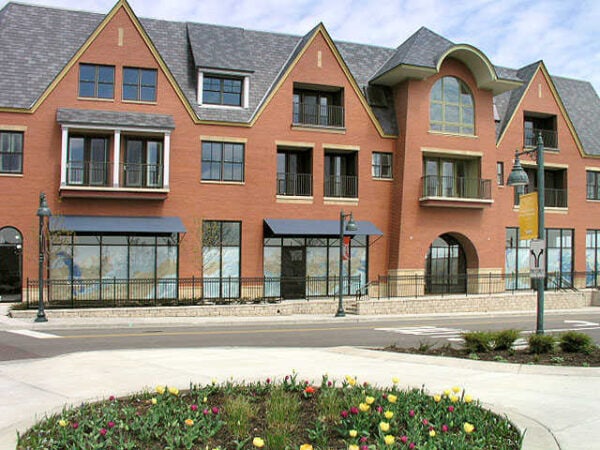 A modern brick building with large windows, upper-floor balconies, and retail spaces on the ground level, facing a street with a landscaped flower bed in front.
