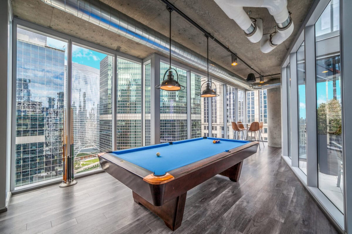 A modern room with a blue pool table, large floor-to-ceiling windows, city skyscraper views, and a small table with chairs in the corner.