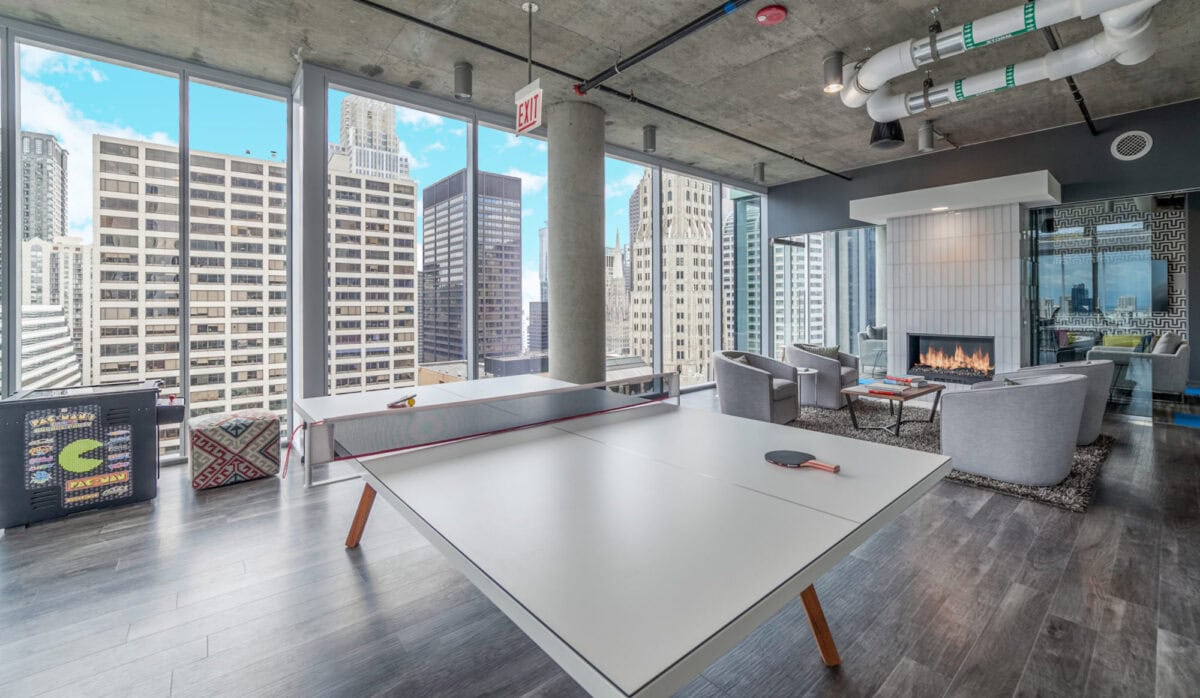 Modern lounge with a ping pong table, fireplace, and seating area, surrounded by large windows overlooking city skyscrapers.