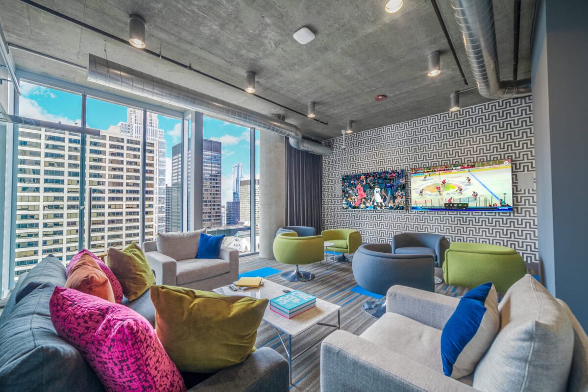 Modern lounge with large windows, colorful chairs, two TVs on a patterned wall, and city buildings visible outside.