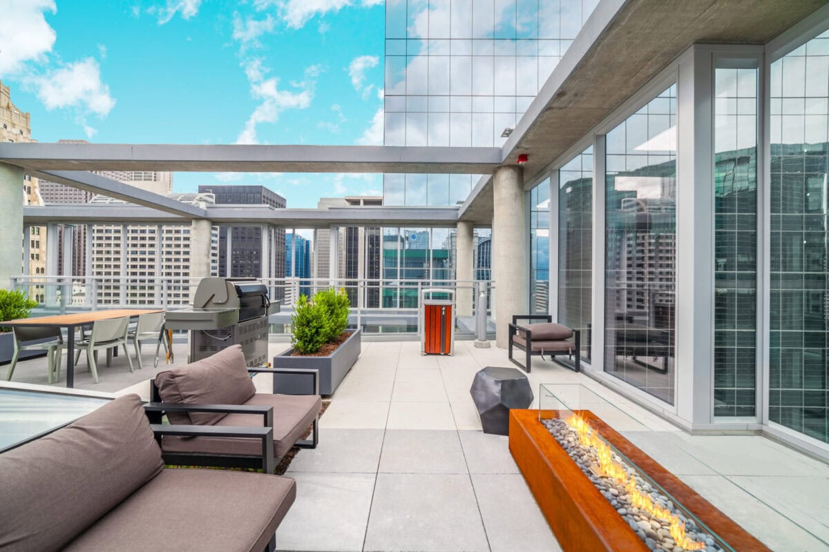 Modern rooftop patio with outdoor seating, gas fire pit, barbecue grill, and city skyscrapers in the background under a partly cloudy sky.