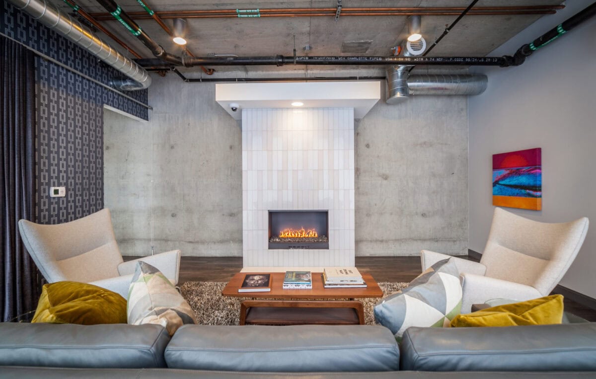 Modern living room with exposed ceiling pipes, concrete walls, a white tiled fireplace, four armchairs, a coffee table, and a sofa with colorful pillows. Abstract art hangs on the wall.