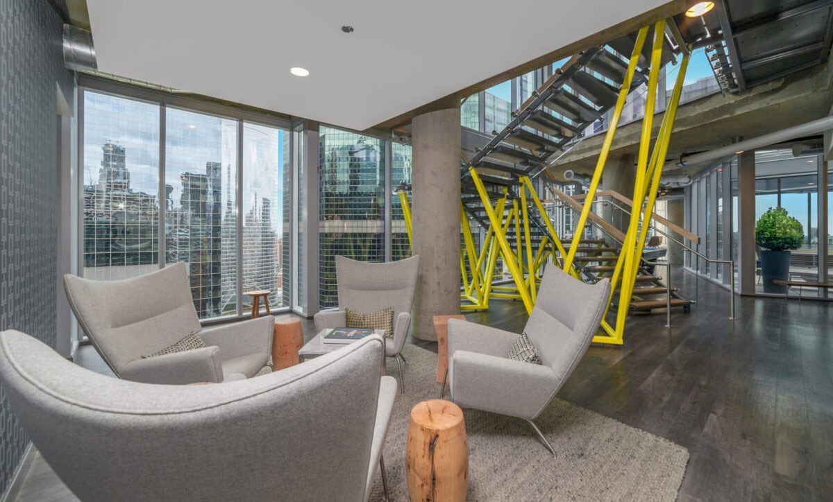 Modern office lounge area with four gray chairs, small wooden tables, and large windows overlooking city skyscrapers; metal staircase with yellow supports in the background.