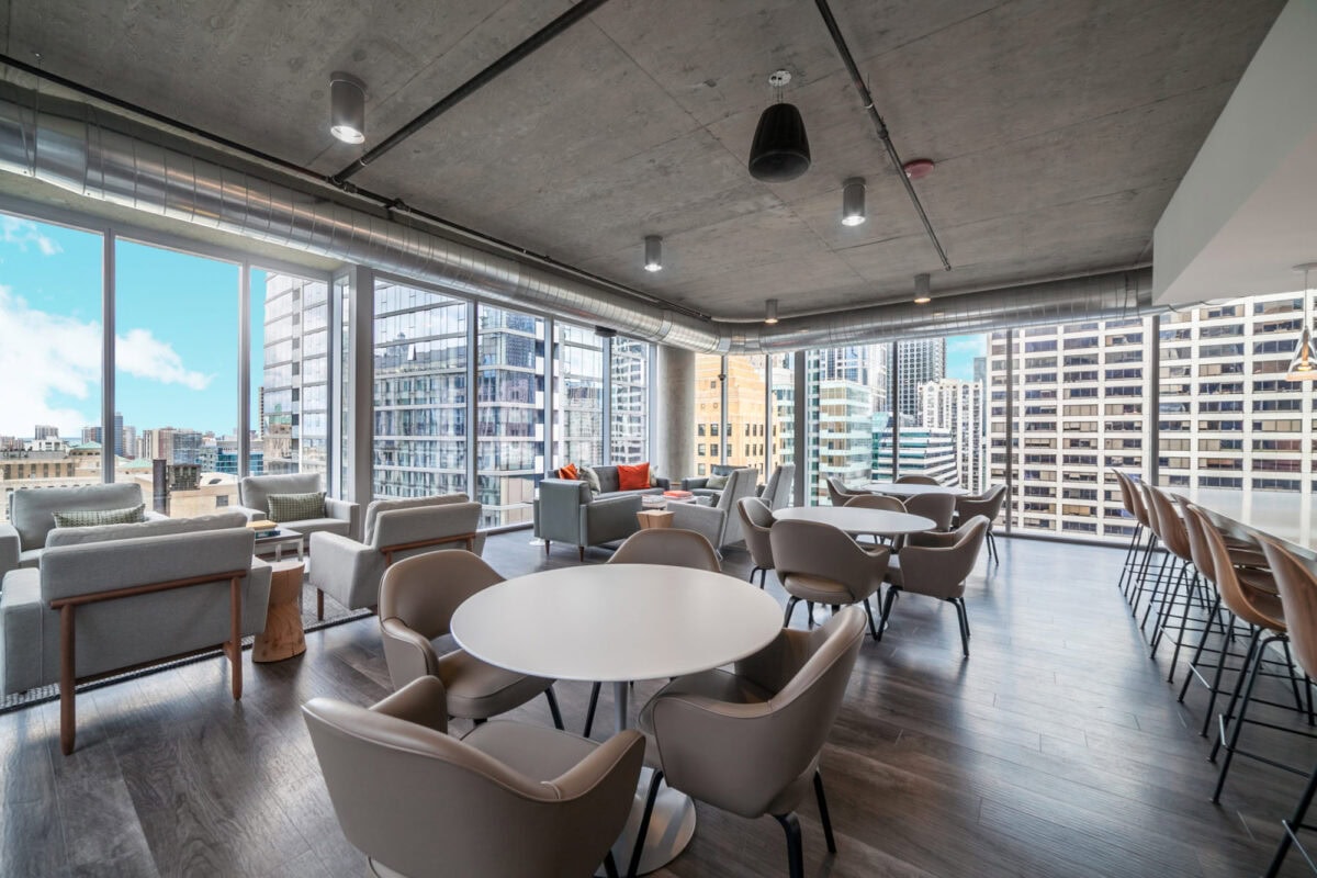 Modern office lounge with round tables, armchairs, bar seating, large windows, and cityscape views under a concrete ceiling.