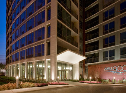 A modern high-rise apartment building with glass windows, a covered entrance, and landscaping, featuring a sign that reads 