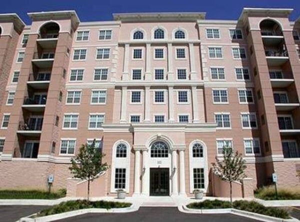 A multi-story pink and white apartment building with balconies, arched entryway, multiple windows, and landscaping in front.