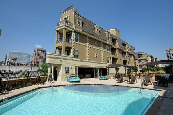 Outdoor swimming pool with lounge chairs and umbrellas in front of a multi-story apartment building on a clear, sunny day.