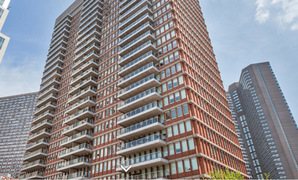 A tall, modern red-brick apartment building with numerous balconies, set against a blue sky, with other high-rise buildings in the background.
