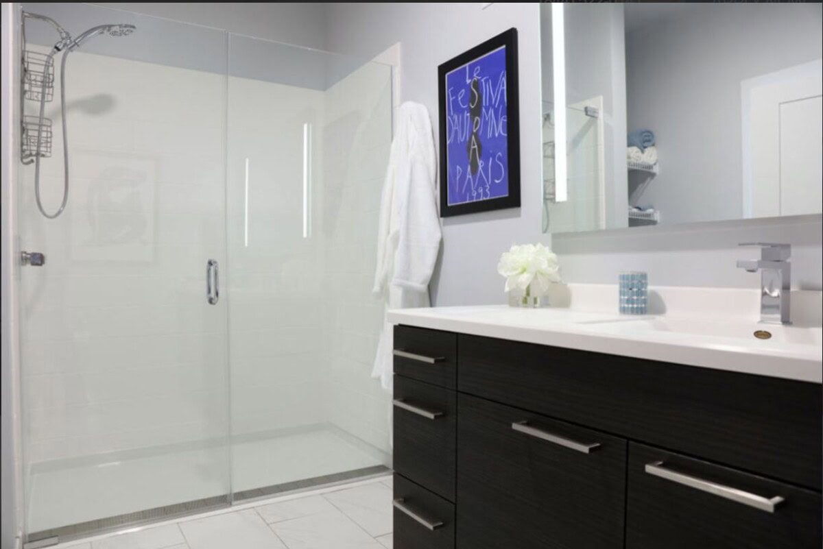 Modern bathroom with a glass shower, dark wood vanity, white countertop, wall art, and a white robe hanging beside the shower.