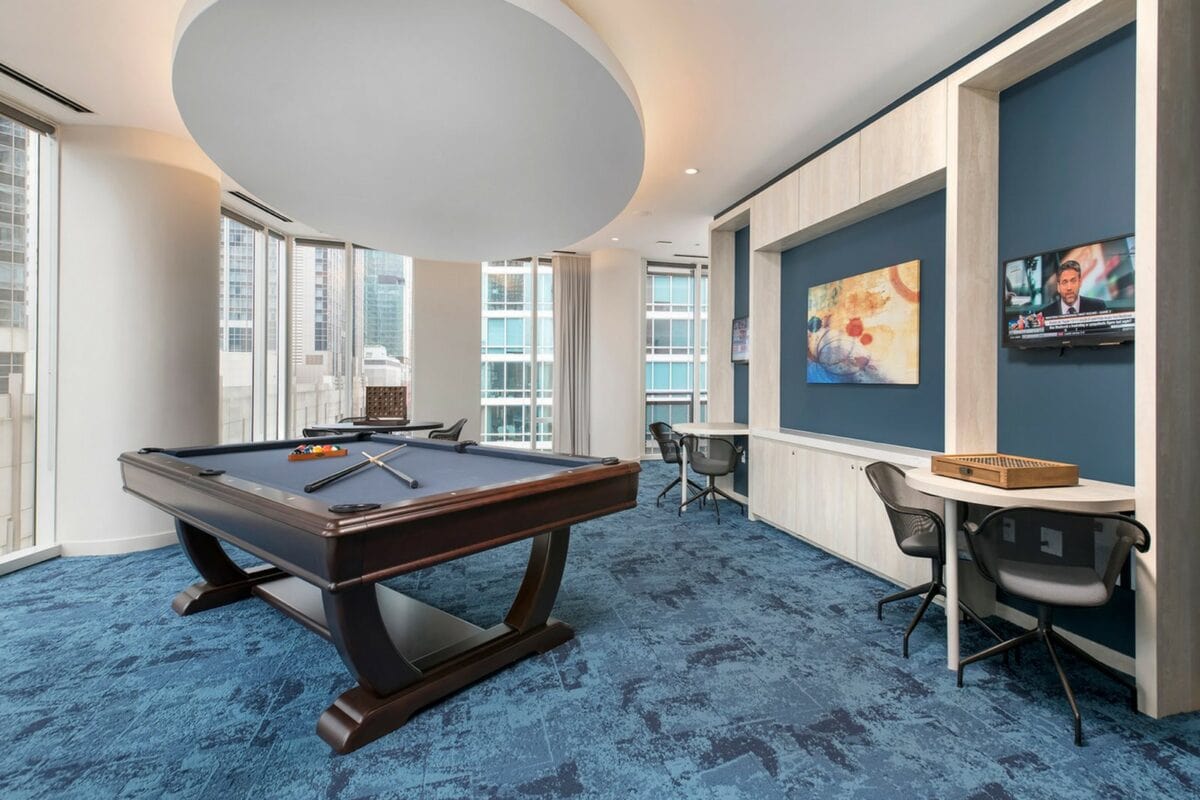 A modern game room with a pool table, two desks with chairs, wall art, a mounted TV, and large windows showing city buildings outside.