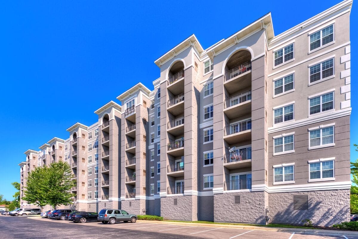 A modern multi-story apartment building with balconies, large windows, and a parking lot with several cars in front under a clear blue sky.