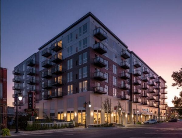 A modern mid-rise apartment building with balconies and ground-floor shops is pictured at dusk, with lights on inside and a clear evening sky in the background.