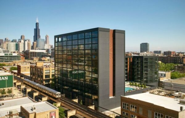 A modern high-rise apartment building stands next to elevated train tracks in an urban area, with the Chicago skyline, including Willis Tower, visible in the background.