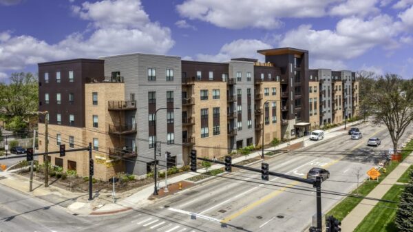 Four-story modern apartment building with brick and dark paneling, balconies, and large windows along a busy intersection under a partly cloudy sky.
