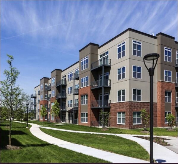 Modern apartment building with multiple floors, large windows, and balconies, surrounded by green lawns, young trees, and a winding sidewalk under a partly cloudy sky.