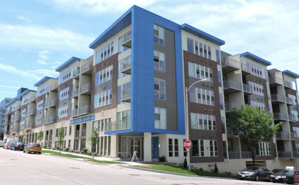 Modern four-story apartment building with blue accents, large windows, multiple balconies, and a stop sign at the street corner on a sunny day.