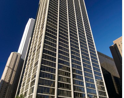Tall modern office building with a grid pattern of windows, set against a clear blue sky with other skyscrapers visible in the background.