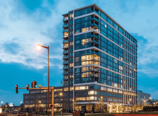A modern high-rise apartment building with glass windows and balconies, illuminated at dusk near a street intersection with traffic lights.