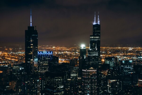 Nighttime cityscape with illuminated skyscrapers and city lights; two tall buildings with glowing spires stand prominently against a dark sky.