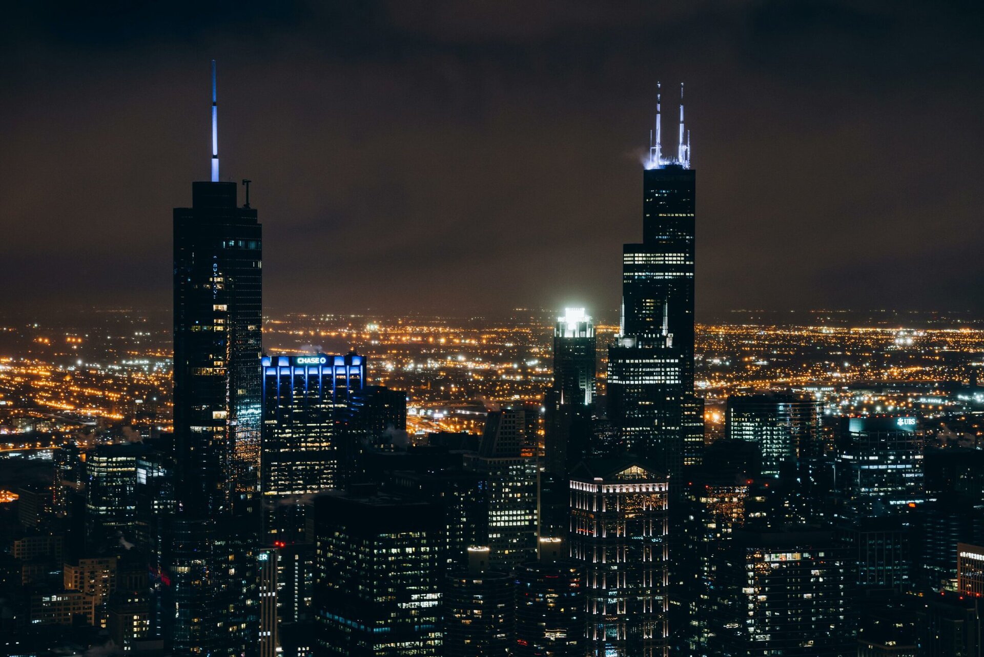 Nighttime cityscape with illuminated skyscrapers and city lights; two tall buildings with glowing spires stand prominently against a dark sky.