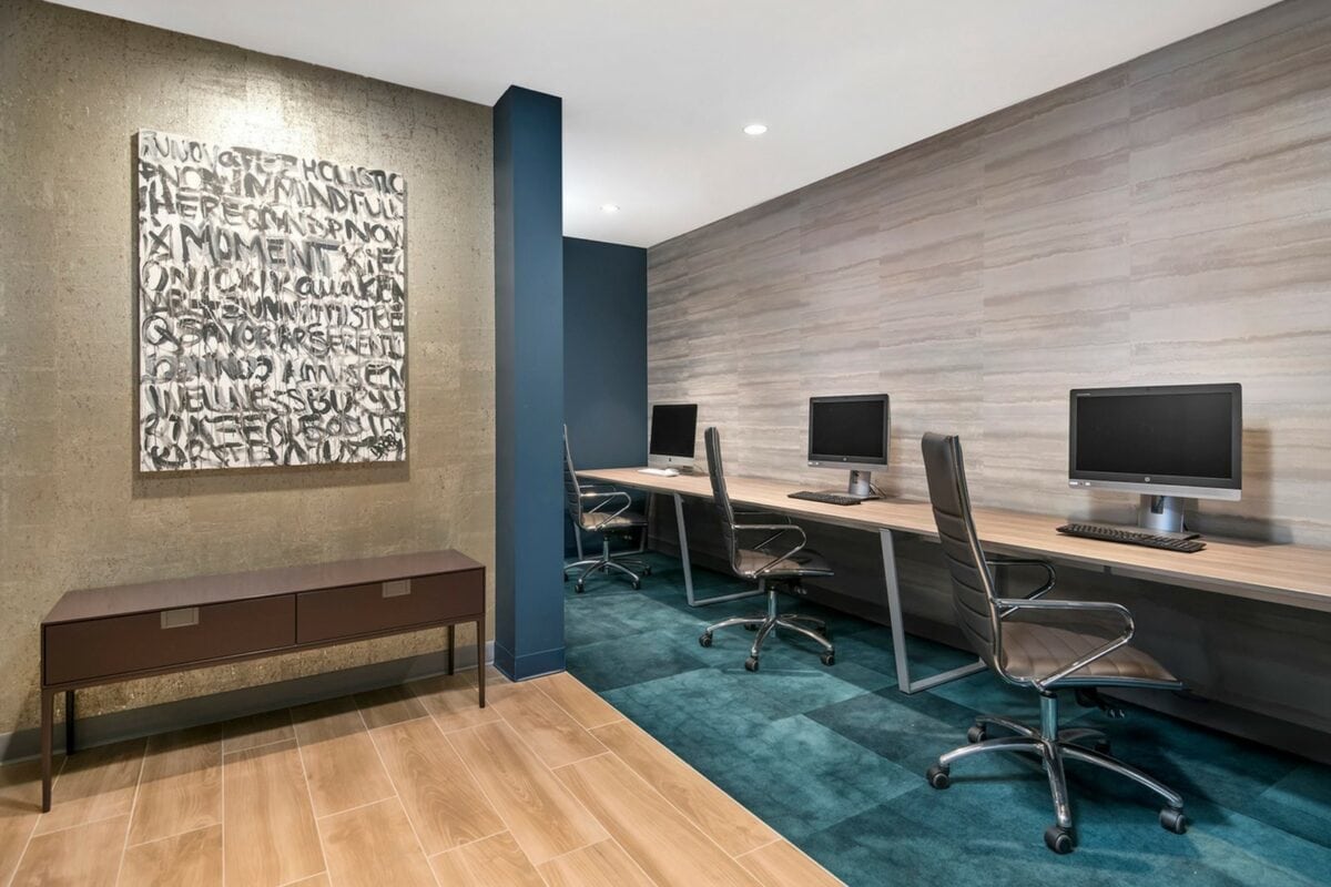 A modern office space with three computer workstations, rolling chairs, a blue accent wall, and an abstract art piece hanging above a brown console table.