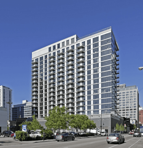 Modern high-rise apartment building with multiple balconies, large glass windows, and street-level retail, surrounded by trees and parked cars on a sunny day.