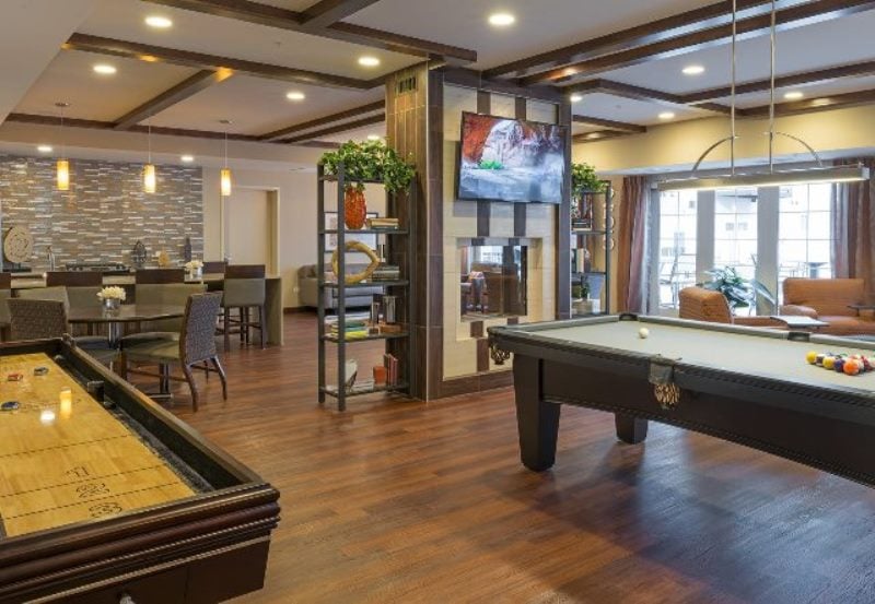 Modern game room with a pool table, shuffleboard, seating area, wall-mounted TV, and wooden flooring.