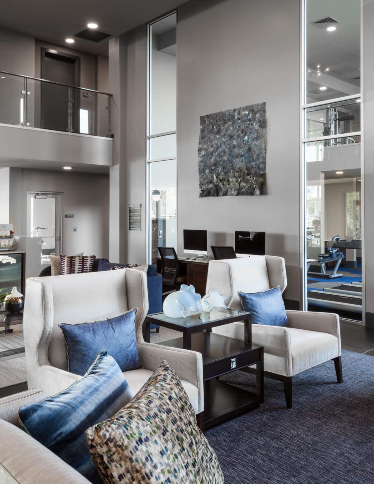 Modern lounge area with armchairs, blue accent pillows, a small table, two computer workstations, large windows, and wall art; glimpses of a gym in the background.
