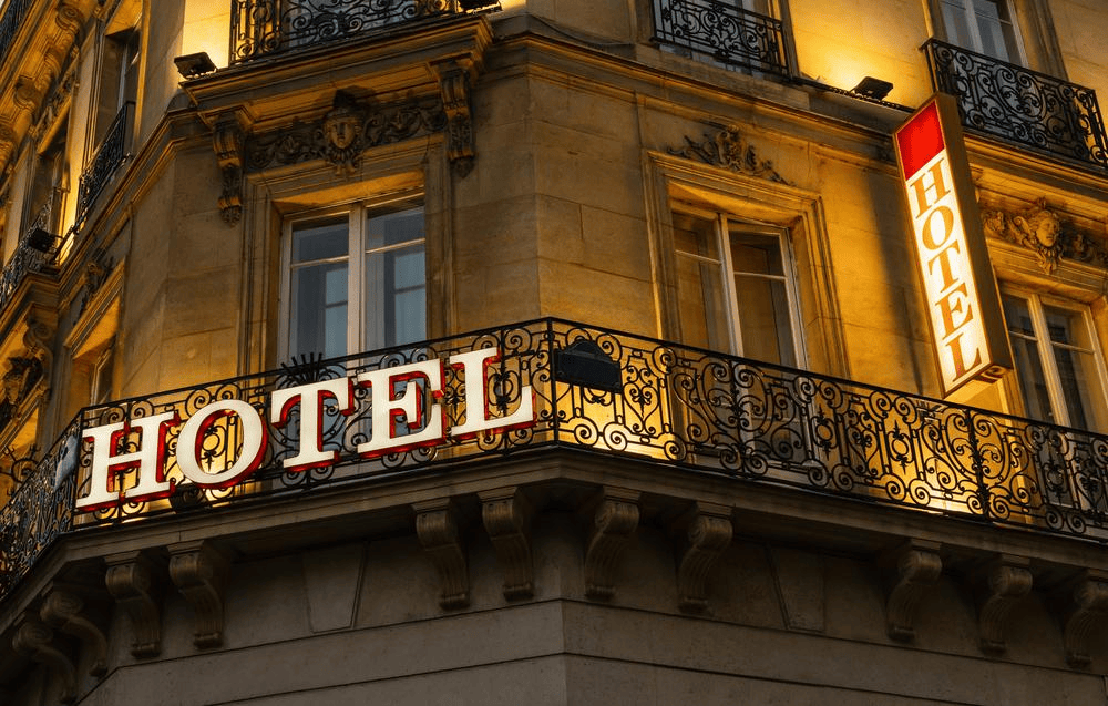 A brightly lit "HOTEL" sign is mounted on the ornate wrought iron balcony of a stone building with large windows.