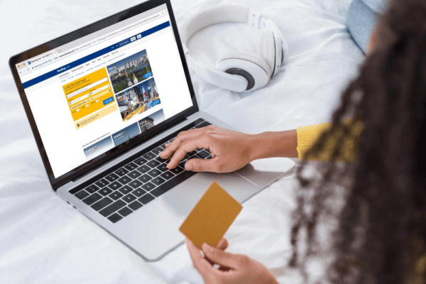 Person holding a credit card while booking a hotel room online on a laptop, with white headphones placed on the bed nearby.
