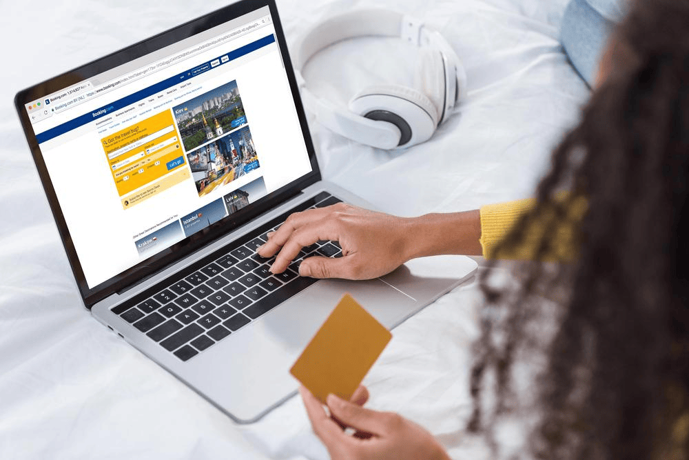 Person holding a credit card while booking a hotel room online on a laptop, with white headphones placed on the bed nearby.