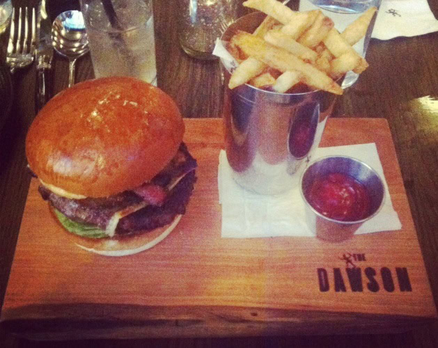 A burger with lettuce and tomato, a cup of french fries, and a small container of ketchup served on a wooden board labeled "THE DANSON.