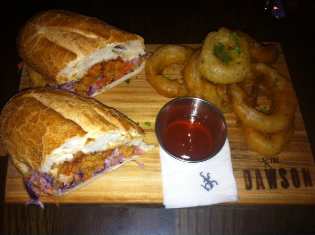 A sandwich with shredded vegetables is served on a wooden board with a side of onion rings and a cup of dipping sauce. The board is branded with "THE DAWSON.
