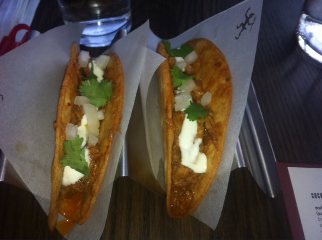 Two tacos in crispy shells, filled with meat, chopped onions, cilantro, and a drizzle of white sauce, served on parchment paper in a metal taco holder.