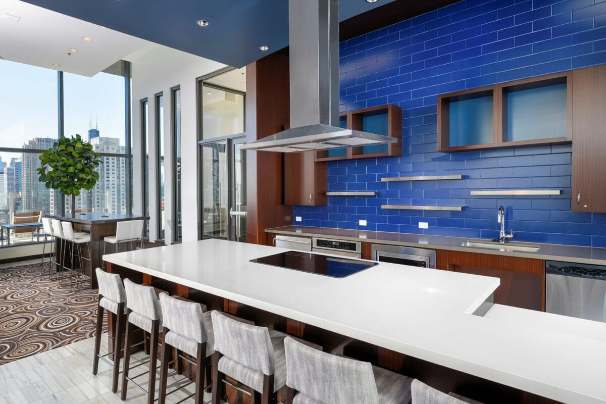 Modern kitchen with a large white island, gray barstools, stainless steel appliances, and a blue tiled backsplash. Floor-to-ceiling windows reveal a cityscape view.